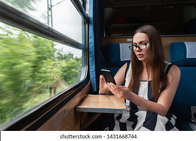 Beautiful Young Brunette Woman In Glasses In A Striped Long Dress Sits By The Window In The Train And Looks Into The Phone. Attractive Girl Cock Over Europe.