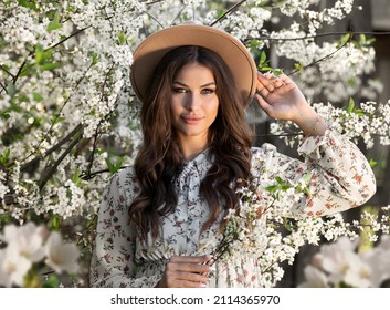 beautiful young brunette woman in a hat with a bouquet of cherry branches in the summer garden, enjoying the blooming spring nature in a sunny day