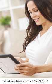 Beautiful Young Brunette Woman At Home Sitting On Sofa Or Settee Using Her Tablet Computer Or IPad And Smiling