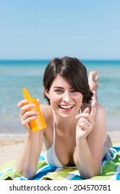 Beautiful Young Brunette Woman Lying On Her Stomach On A Towel At The Beach Applying Sunscreen Smiling Playfully As She Holds Up A Blob Of The Lotion On Her Finger For The Camera