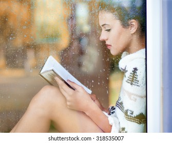 Beautiful Young  Brunette Woman Reading Book Wearing Knitted Dress Sitting Home Behind A Window Covered With Rain Drops. Blurred Fall Garden Reflection On The Glass. Raining Autumn Concept