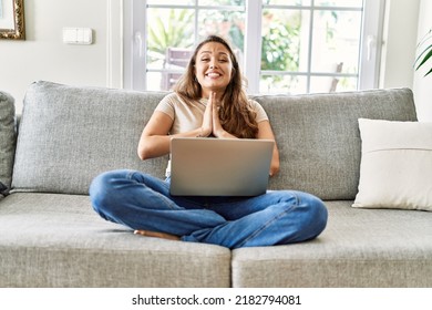 Beautiful Young Brunette Woman Sitting On The Sofa Using Computer Laptop At Home Praying With Hands Together Asking For Forgiveness Smiling Confident. 