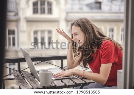 Beautiful young brunette woman sitting on balcony using laptop while having online chat consultation