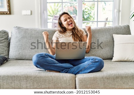 Beautiful young brunette woman sitting on the sofa using computer laptop at home very happy and excited doing winner gesture with arms raised, smiling and screaming for success. celebration concept.  Foto d'archivio © 