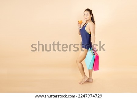 Beautiful young brunette woman slim body wearing blue one-piece swimsuit hold glass of orange dink and shopping bags in hand isolated on beige background, studio shot. Summer holidays concept.