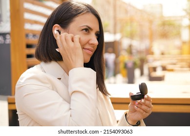 The Beautiful Young Brunette Woman Takes Out A Wireless Earpiece From A Portable Charging Box And Inserts It Into Her Ear. Black Earbuds. Listen To Music Anywhere And Enjoy The Sound