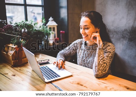 beautiful young brunette woman using a laptop at coffee shop at a wooden table near window typing text on a keyboard. In winter, with the light from the lamp, she is dressed in a warm gray sweater.