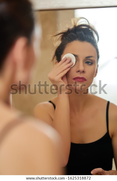 Beautiful young brunette woman washes away makeup\
in front of bathroom\
mirror