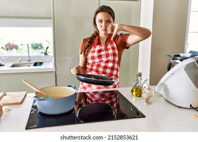 Beautiful Young Brunette Woman Wearing Apron Cooking At The Kitchen With Angry Face, Negative Sign Showing Dislike With Thumbs Down, Rejection Concept 