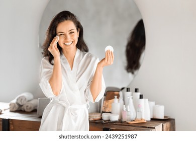 Beautiful young brunette woman wearing white silky bathrobe standing next to vanity table with mirror above, using cotton pads, cleansing face, removing makeup, moisturizing skin in bathroom - Powered by Shutterstock