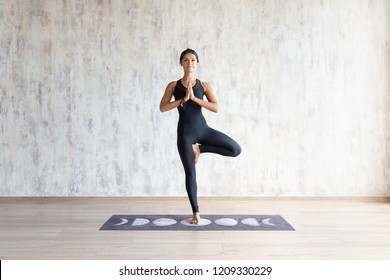 Beautiful Young Brunette Woman Yoga Instructor Doing Vrikshasana On A Mat In A Wooden Floor Standing In The Gym With Day Lighting