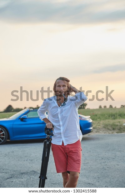 The\
beautiful young brutal man stands at a blue sports car at sunset,\
he is dressed in a white shirt with a short sleeve and red shorts,\
the photographer stands leans on a tripod, he\
smiles