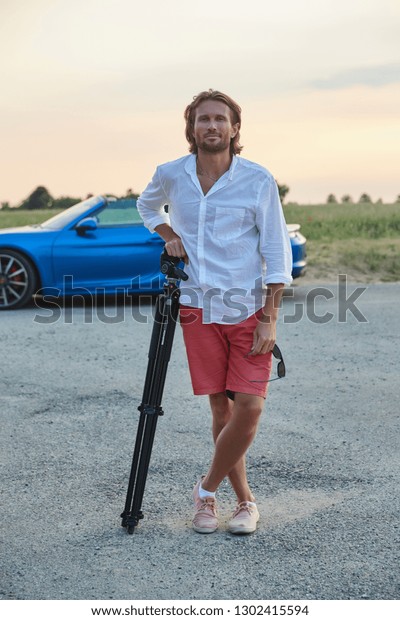 The\
beautiful young brutal man stands at a blue sports car at sunset,\
he is dressed in a white shirt with a short sleeve and red shorts,\
the photographer stands leans on a tripod, he\
smiles
