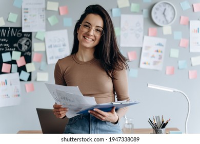 Beautiful, young business lady holds important documents in her hands, she reviews them. Smiling, charming girl with glasses works in a modern office. - Powered by Shutterstock