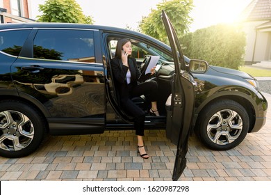 Beautiful Young Business Woman In Black Suit Using Mobile Phone And Making Notes On Papers, While Sitting In The Car With Open Door