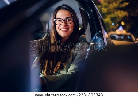 Beautiful young business woman driving a car while traveling during a night and looking into the camera and smiling. Gorgeous female driving a vehicle in the city during night. Copy space.