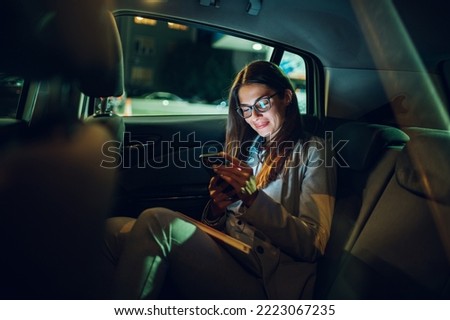 Beautiful young business woman smiling and use smartphone inside the car while traveling during a night. Contacting friends or business associates when you are away. Typing a message or an email.