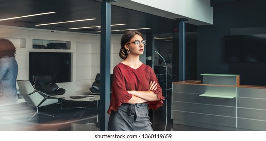 Beautiful Young Business Woman Standing In Lobby Of An Office With Her Arms Crossed, Looking Ahead, With People Walking Past In A Blur