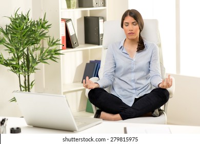 Beautiful Young Businesswoman Practicing Yoga In The Office. She Sits On Lotus Pose In A Chair In Front Of Her Desk.