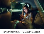 Beautiful young businesswoman traveling with car on a business trip during night while sitting in a back seat and using a smartphone. Gorgeous female using mobile phone to send email or messages.