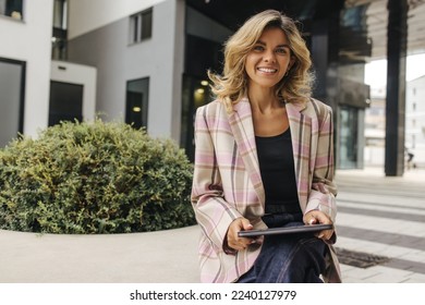 Beautiful young caucasian business woman working on tablet near office. Blonde smiles looking at camera, wears jacket. Concept of use - Shutterstock ID 2240127979