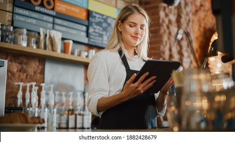 Beautiful Young Caucasian Coffee Shop Manager with Blond Hair is Making Notes on a Tablet Computer and Orders Inventory Items for the Menu in a Cozy Loft-Style Cafe. Successful Restaurant Owner.