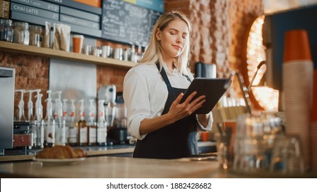 Beautiful Young Caucasian Coffee Shop Manager with Blond Hair is Making Notes on a Tablet Computer and Orders Inventory Items for the Menu in a Cozy Loft-Style Cafe. Successful Restaurant Owner. - Powered by Shutterstock