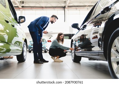 Beautiful young caucasian female client customer choosing new car, trying checking its options, tire, wheels while male shop assistant helping her to choose it at dealer auto shop