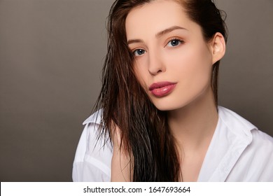 A Beautiful Young Caucasian Girl With Blue Eyes, With Long Dark Wet Loose Hair In A White Shirt Looks At The Camera. Close Up Portrait On A Dark Background.