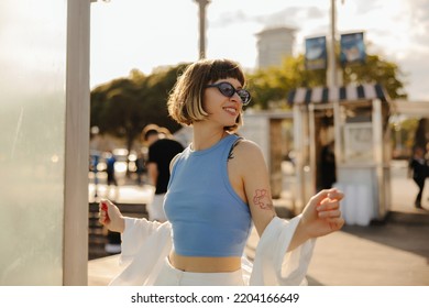 Beautiful young caucasian girl with brown hair with tattoo walks in summer city center. Model wears sunglasses, blue tank top and white shirt. People sincere emotions lifestyle concept.