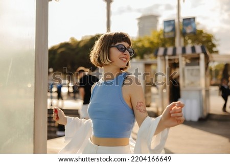 Beautiful young caucasian girl with brown hair with tattoo walks in summer city center. Model wears sunglasses, blue tank top and white shirt. People sincere emotions lifestyle concept. Stock photo © 