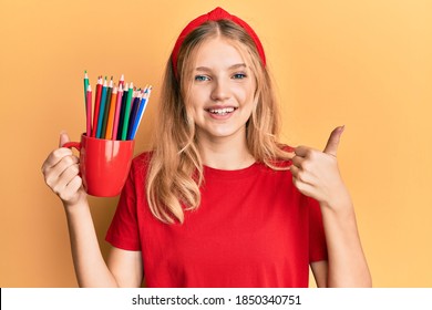 Beautiful young caucasian girl holding art colored pencils smiling happy and positive, thumb up doing excellent and approval sign  - Powered by Shutterstock