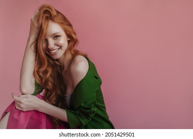 Beautiful young caucasian lady smiling shyly while sitting on pink background. Redhead, dressed in silk shirt and fuchsia skirt, lifts her wavy hair with her hand and reveals bare shoulder.