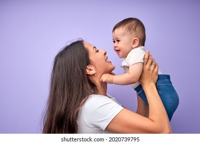 Beautiful Young Caucasian Mother Tossing Up Child Baby. Baby Laughing. Purple Background. In Studio. Copy Space, Side View. Portrait Of Caucasian Family. Happy Childhood Concept