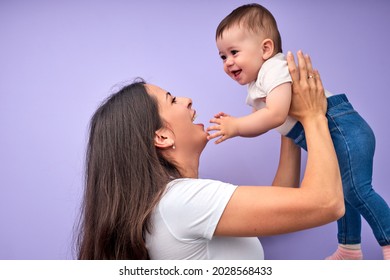 Beautiful Young Caucasian Mother Tossing Up Child Baby. Baby Laughing. Purple Background. In Studio. Copy Space, Side View. Portrait Of Caucasian Family. Happy Childhood Concept