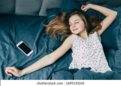 A Beautiful Young Caucasian Teenage Girl With Long Hair Is Lying Next To The Phone Stretching In Bed And Has Not Yet Woken Up. Teenager Fell Asleep Because Of Late Chat With Friends. Lazy Morning