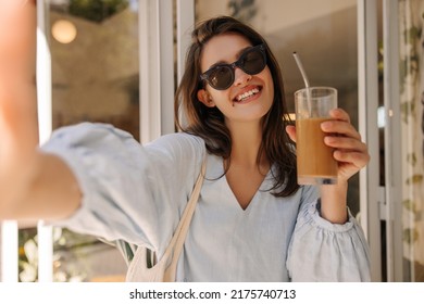 Beautiful Young Caucasian Woman With Chocolate Smoothie Takes Selfie Outdoors. Brunette Wears Sunglasses And Blue Dress In Summer. Lifestyle Concept