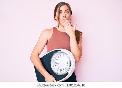 Beautiful Young Caucasian Woman Holding Weight Machine To Balance Weight Loss Covering Mouth With Hand, Shocked And Afraid For Mistake. Surprised Expression 