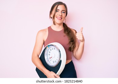 Beautiful Young Caucasian Woman Holding Weight Machine To Balance Weight Loss Smiling Happy And Positive, Thumb Up Doing Excellent And Approval Sign 