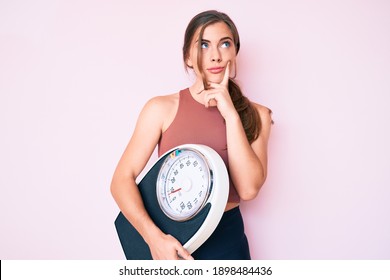 Beautiful Young Caucasian Woman Holding Weight Machine To Balance Weight Loss Serious Face Thinking About Question With Hand On Chin, Thoughtful About Confusing Idea 