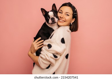Beautiful young caucasian woman hugging her pet boston terrier breed on pink background. Brunette presses dog to while holding it in her arms. Animal love concept