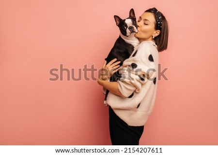 Beautiful young caucasian woman kissing her cute dog on pink background with space for text. Brunette girl in white sweater shares love with pet. Showing affection concept