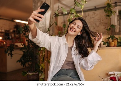 Beautiful Young Caucasian Woman Is Photographed On Smartphone Sitting Indoors During Day. Brunette With Snow-white Smile Wears Casual Clothes. Technology Concept