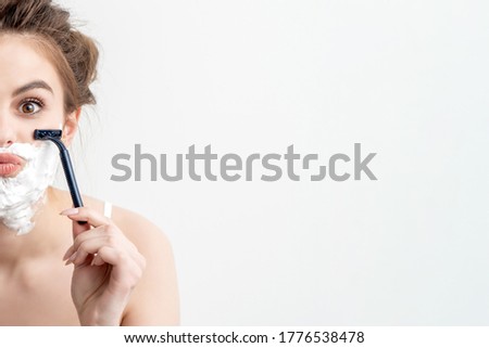 Beautiful young caucasian woman shaving her face by two razors on white background. Pretty woman with shaving foam on her face