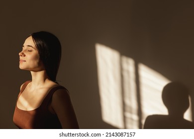 Beautiful Young Caucasian Woman In Sunlight With Closed Eyes Sees Off Sunset Indoors. Smiling Fashion Model Wears Sporty Brown Top. Enjoying Little Things, Positive Attitude Concept