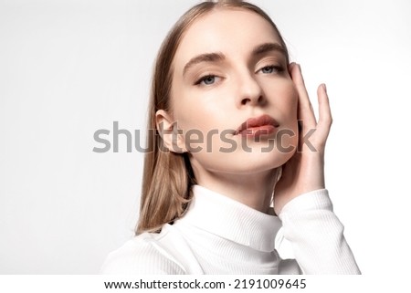 Beautiful young caucasian woman touching her smooth soft facial skin. Model with natural makeup wearing white turtleneck looking to camera. Advertising of skincare treatment cosmetics, beauty products