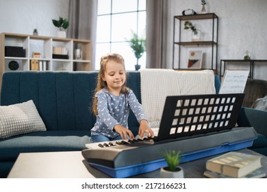 Beautiful young charming little girl smiling while playing classic digital piano at home. Caucasian musician girl studying to play piano indoor in light living room.