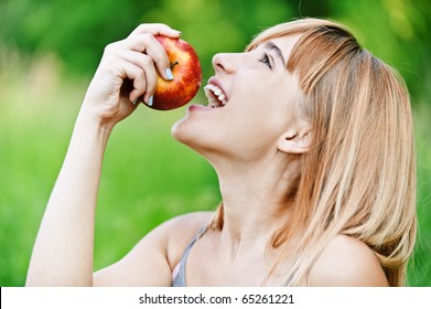 Beautiful young cheerful woman has turned in profile and bites red big apple, against green summer garden.