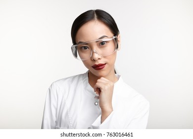 Beautiful young Chinese woman laboratory worker wearing red lipstick, protective goggles and white medical gown holding hand on her chin and smiling while working, conducting scientific research