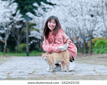 Beautiful young Chinese woman playing with homeless ginger tabby cat with blurry blooming cherry tree background. Candid happy moment. Emotions, people, beauty, youth and lifestyle portrait.